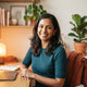 A professional portrait of a smiling South Asian woman in a warm, modern home office setting.