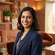 A professional portrait of a South Asian woman smiling in a bright, modern office space with warm amber and terracotta tones.