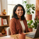 A professional portrait of a South Asian woman smiling in a bright, warm-toned home office environment.